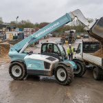 A light-blue Zeromachine telehandler tips a bucket of sand into the back of a flatbed truck in a builders’ merchant yard, with stacked pallets of materials and a covered storage area in the background on an overcast day.